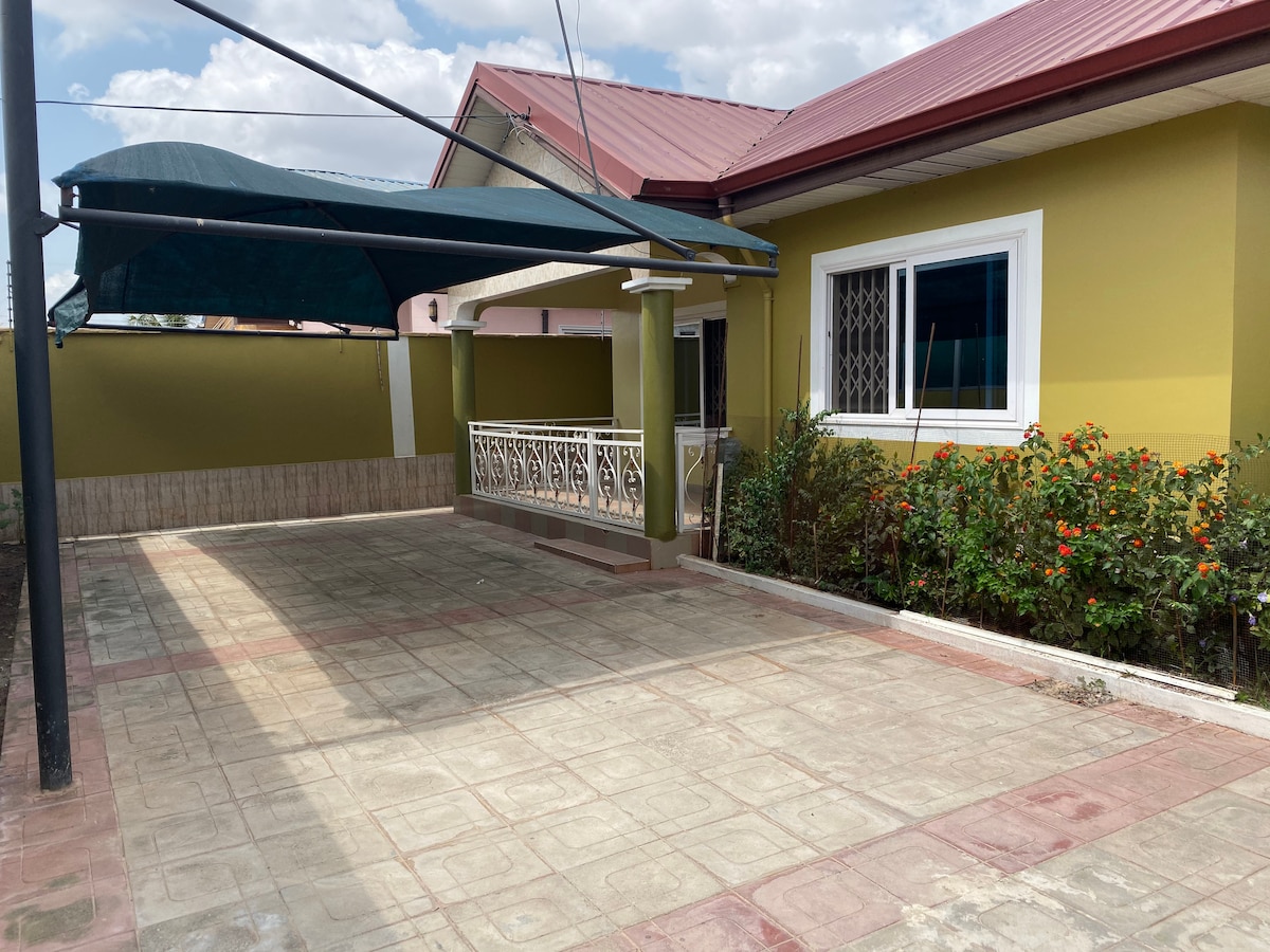 A covered parking area is shown, featuring a concrete floor with a grid pattern. The structure includes a low wall and a roof, providing shade. Colorful plants border the space, adding a natural element to the otherwise paved area.