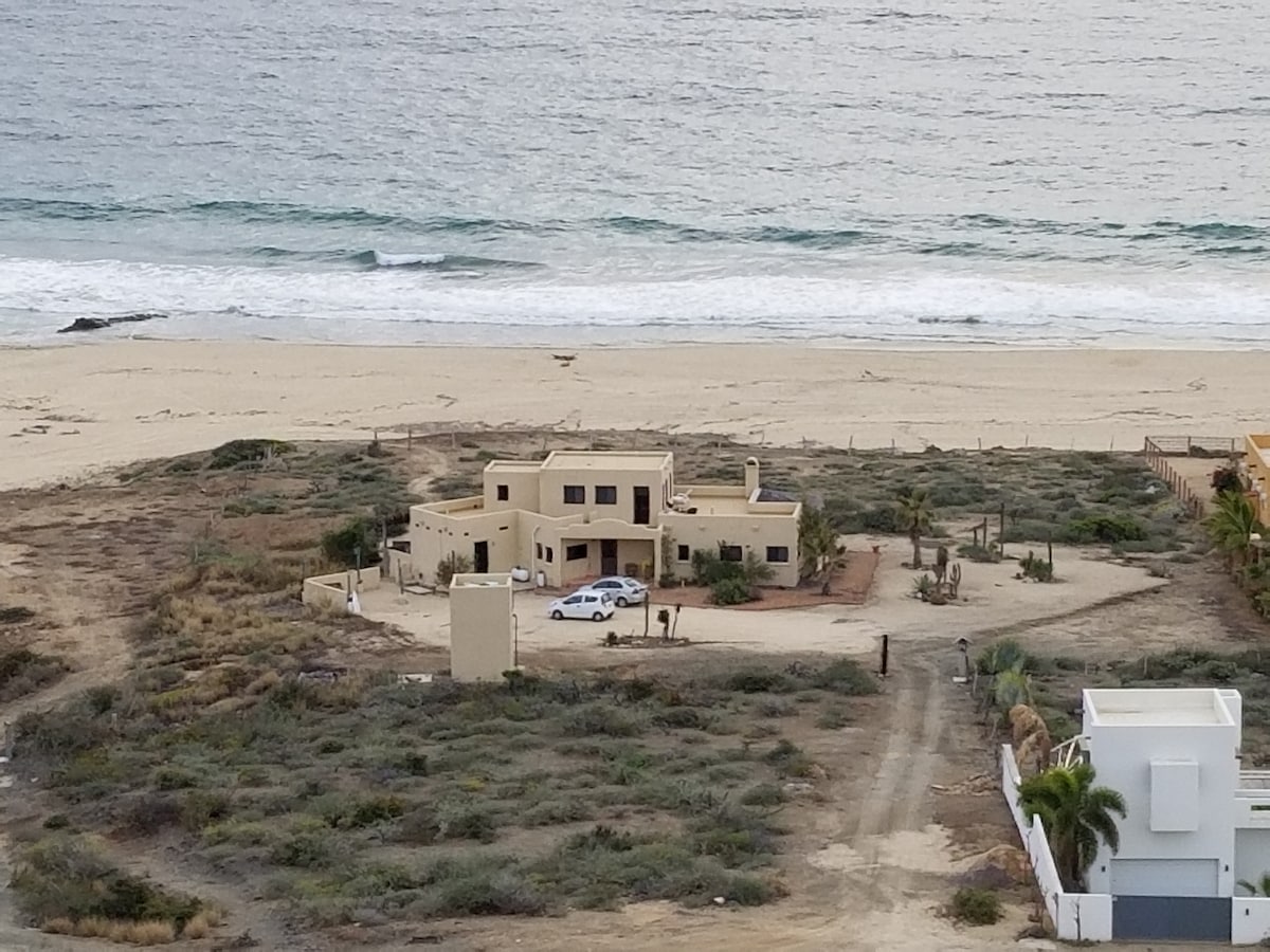 The house is positioned prominently on an expansive stretch of beach, surrounded by natural vegetation. A sandy area leads toward the waves, with the ocean visible in the background. The property features a clean, functional design, with a light exterior and a private driveway.