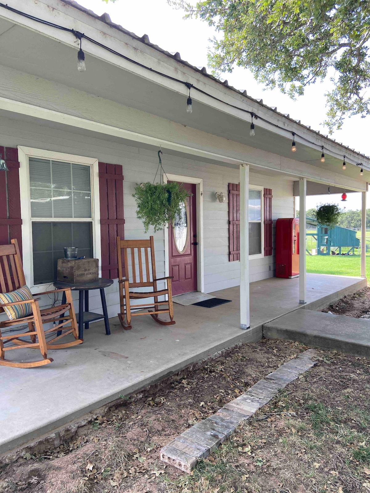 The guest house features a welcoming front porch with two wooden rocking chairs and a small table. Red shutters complement the entrance door. Decorative string lights hang overhead, enhancing the outdoor space, while a grassy area is visible beyond the porch.