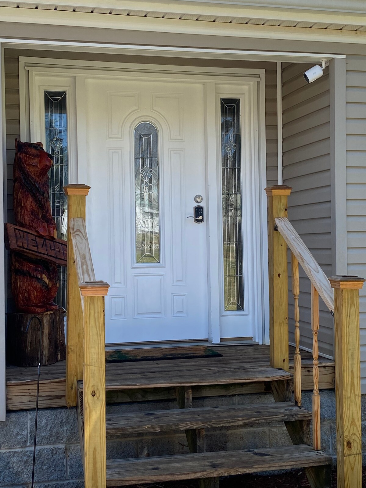 The entryway features a white front door with decorative glass panels. The wooden steps lead up to the door, which is framed by light-colored siding. A carved wooden bear and a sign are positioned nearby, adding character to the welcoming entrance.