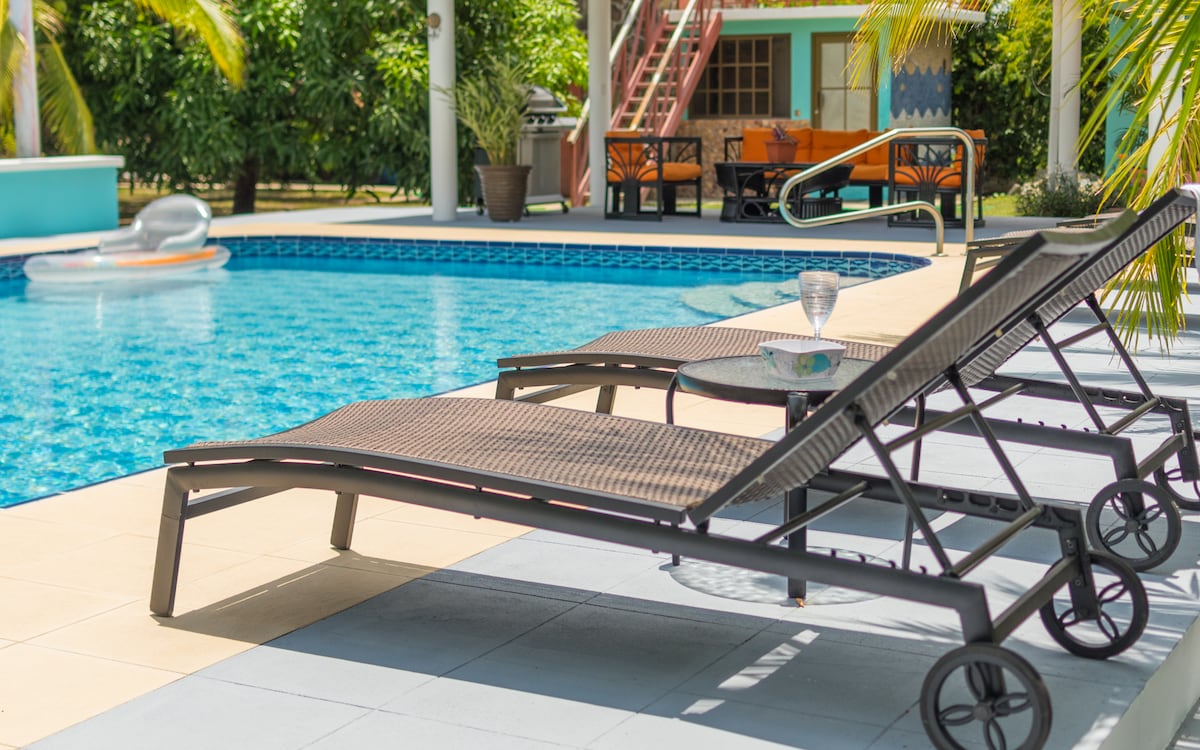 Two lounge chairs are positioned beside a spacious swimming pool, surrounded by lush greenery. A small table between the chairs holds a glass, and the calming blue of the water reflects the sunlight. The area invites relaxation under the tropical setting.