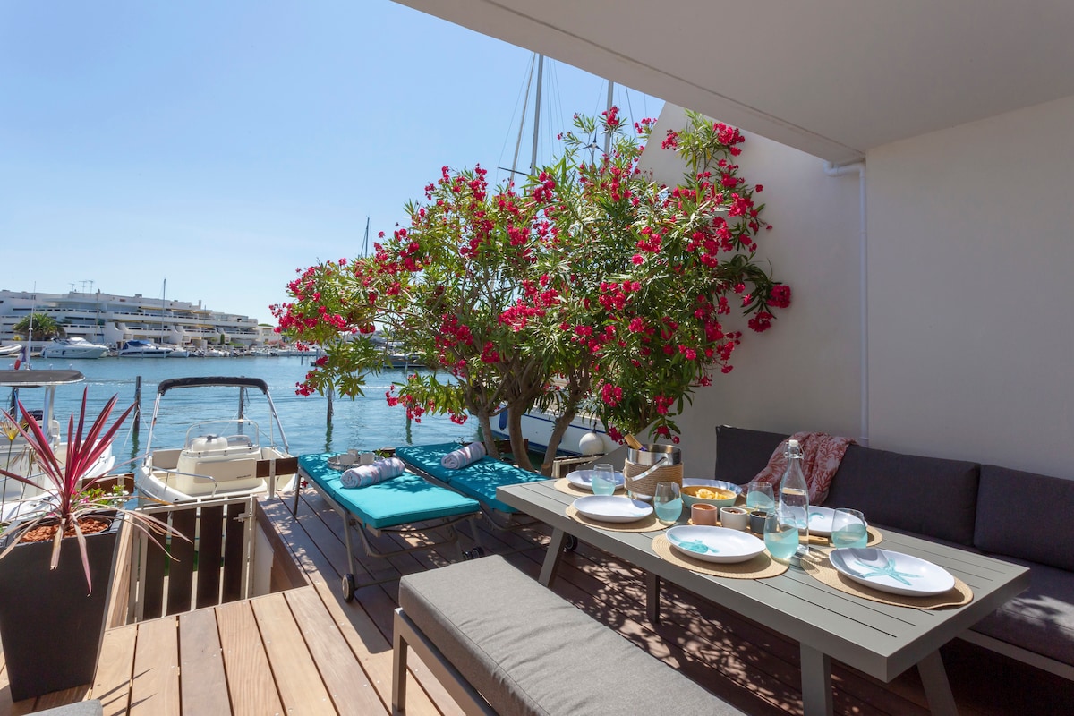 An outdoor terrace is visible, featuring a dining setup with tableware neatly arranged. Comfortable seating is surrounded by vibrant flowering plants, enhancing the view of the water. Sun loungers invite relaxation, providing a serene atmosphere for enjoying meals or leisure time.