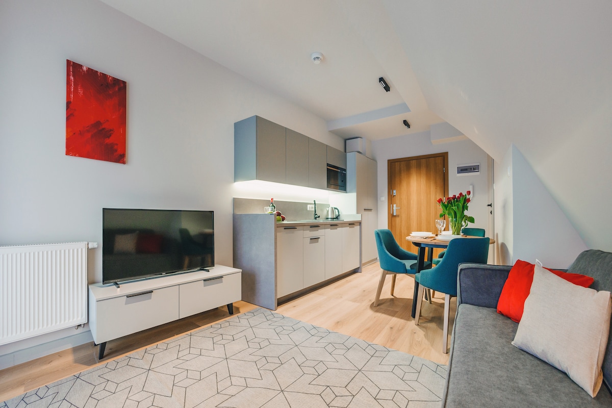 A modern studio apartment features a sleek open layout. A functional kitchen is equipped with essential appliances, adjacent to a cozy seating area with a gray sofa. A television is positioned on a light-colored media unit, while a wooden dining table is set for four, enhancing the inviting ambiance.