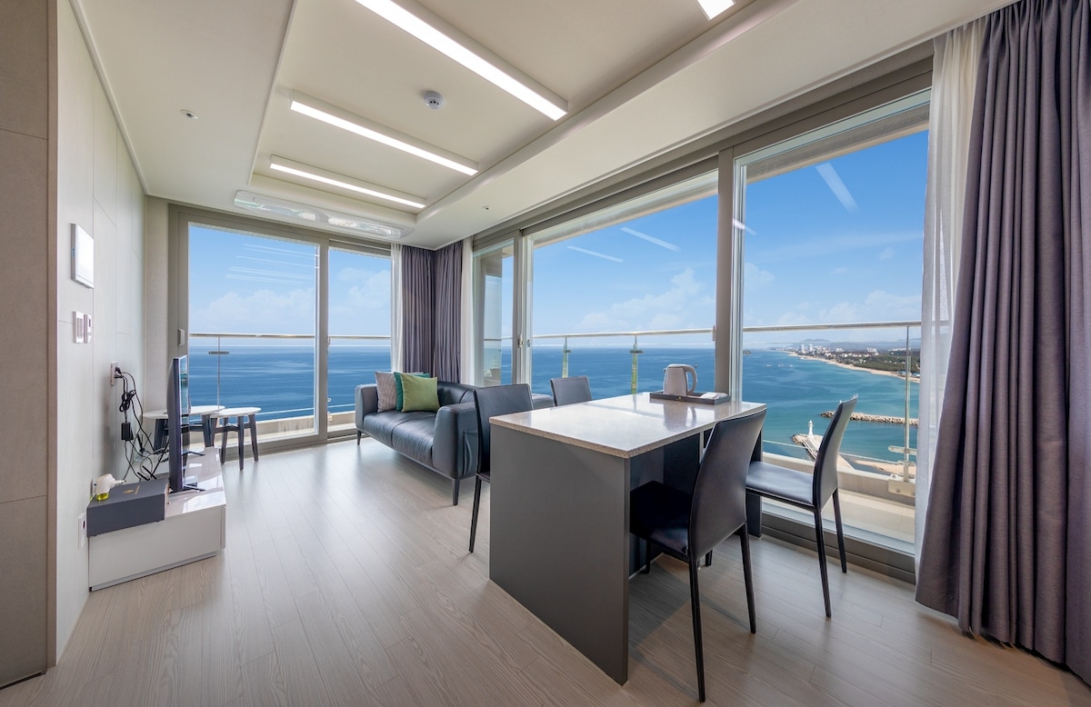A spacious living area is displayed with large windows showcasing panoramic ocean views. A modern black sofa is positioned beside a dining table, creating a welcoming space. Natural light fills the room, enhancing the neutral tones of the decor.