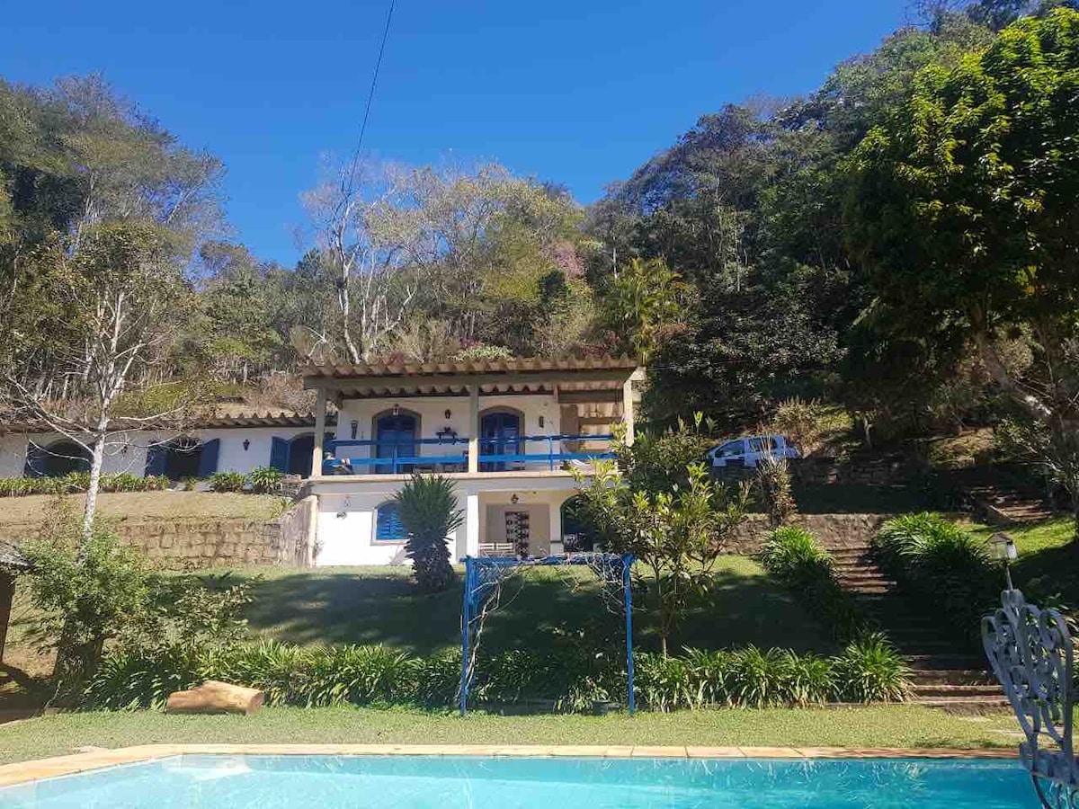 A charming rural property is seen amidst lush greenery, featuring a two-story house with blue accents and a covered porch. The well-maintained garden offers a blend of trees and plants, while a clear pool reflects the bright blue sky.
