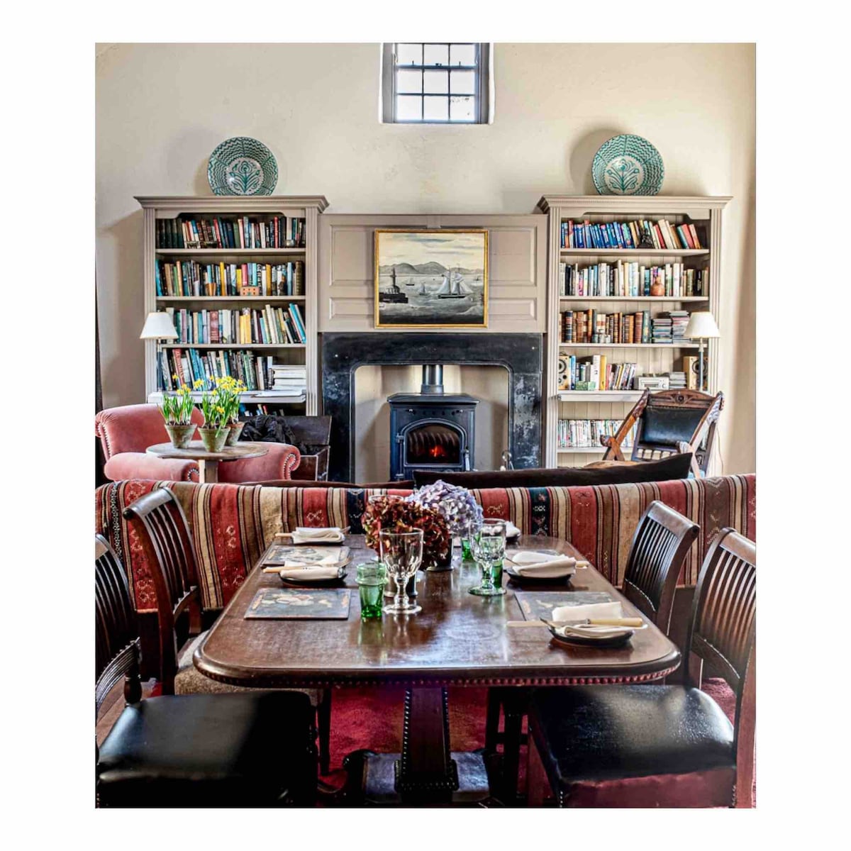 A cozy living area features a dark wood dining table set for a meal. Bookshelves filled with various books frame a traditional fireplace, adding warmth to the space. Comfortable seating options, including a red armchair and decorative plants, create an inviting atmosphere.