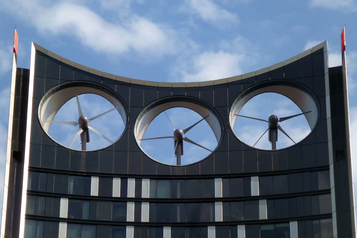 The distinctive rooftop of the Strata Building is highlighted by three large wind turbines. The modern architectural design features vertical lines and a glass facade, with the turbines prominently centered against a backdrop of blue sky and scattered clouds.