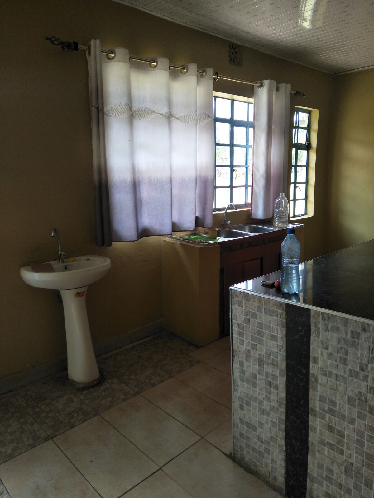 A functional kitchen area features a sink and a countertop with a mosaic design. Natural light enters through two windows dressed with sheer curtains, illuminating the tiled floor. A water bottle sits on the counter, enhancing the space's practicality.