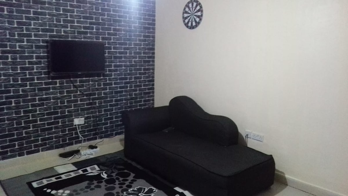 A relaxation area features a dark, curved sofa positioned against a textured black wall. A flat-screen television is mounted above a low power outlet. A circular dartboard adds a recreational element, and a patterned area rug grounds the space.
