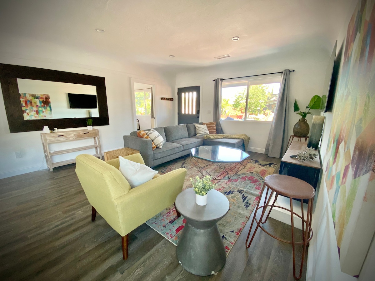 A spacious living area features a large gray sectional sofa, accented by colorful cushions. A round coffee table sits in the center on a patterned rug. A minimalistic side table and a stool are nearby. Natural light filters through windows, illuminating the modern decor.