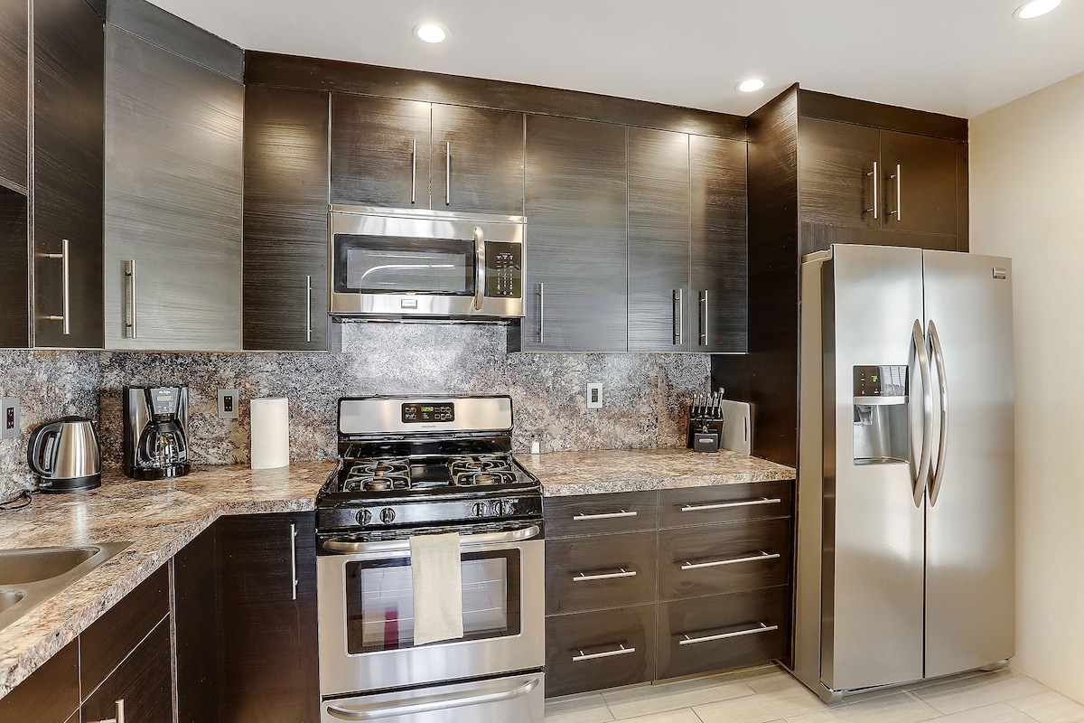 A modern kitchen is equipped with stainless steel appliances, including a gas range and a large refrigerator. The cabinetry is finished in dark tones, complementing the granite countertops. Under-cabinet lighting illuminates the work areas, while a coffee maker and kettle sit on the countertop.