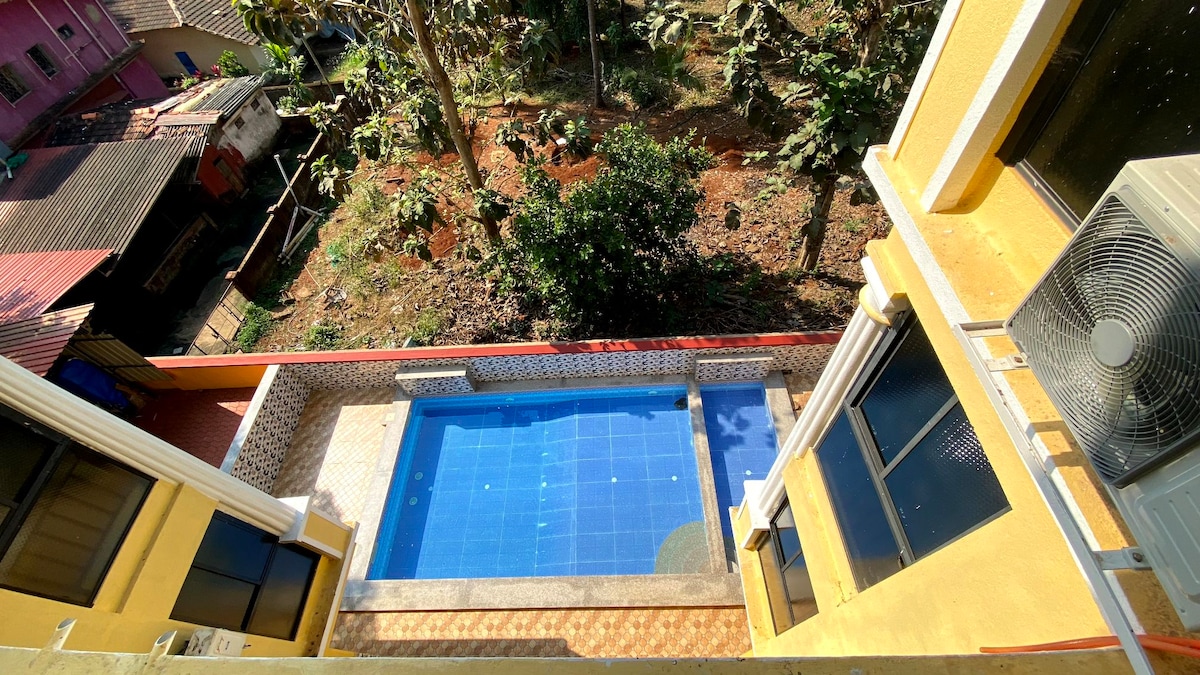 A clear, aerial view of the outdoor swimming pool is showcased, featuring blue tiles and surrounded by a tiled patio. Lush greenery and tropical trees can be seen in the background, providing a serene, natural setting.