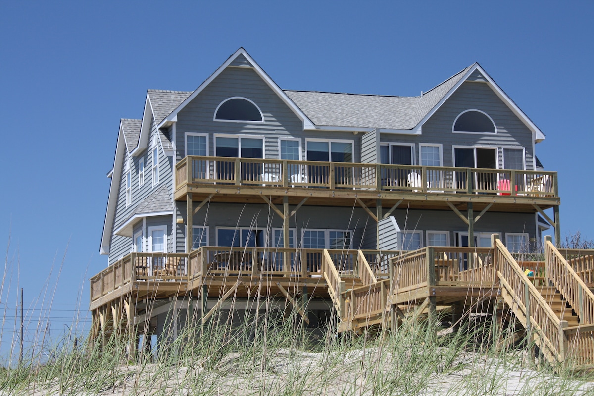 A spacious oceanfront duplex is set against a clear blue sky, featuring multiple decks with wooden railings. The well-maintained exterior is complemented by large windows, allowing for ample natural light. Surrounding foliage adds a subtle touch to the inviting beachside setting.