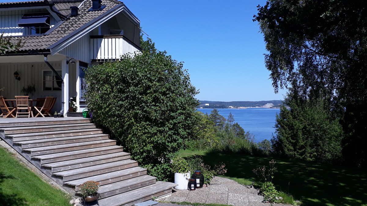 A spacious outdoor area features wooden steps leading down towards a scenic view of Byfjorden. Lush greenery surrounds the space, with a glimpse of the water visible in the distance. Sunlight creates a bright atmosphere, enhancing the calmness of the setting.