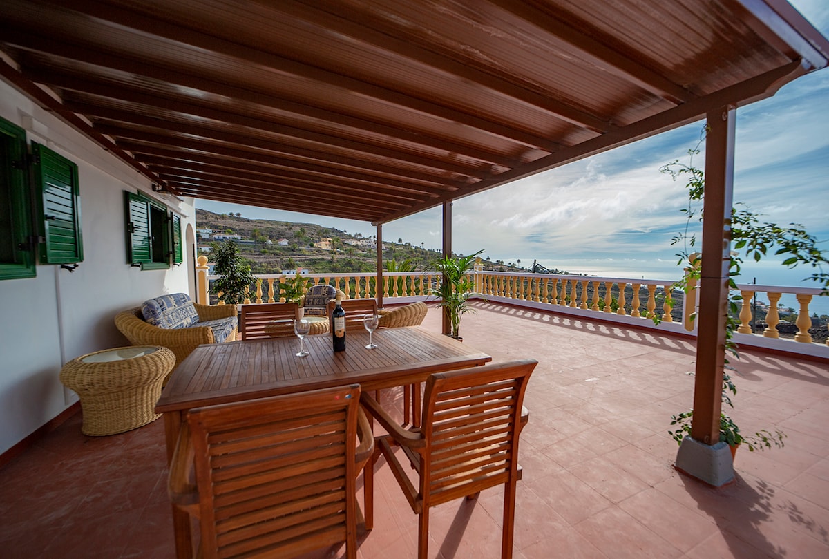 A spacious terrace is covered by a wooden roof, featuring a dining table surrounded by several wooden chairs. A comfortable wicker chair is positioned nearby. Lush greenery and scenic views enhance the outdoor space, while two green-shuttered windows provide natural light.