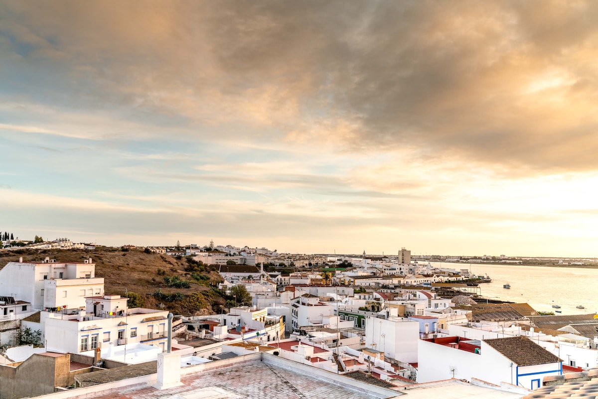 A scenic view of a coastal town, featuring whitewashed buildings that reflect the soft colors of a sunset sky. The landscape includes distant hills and waterways, creating a serene backdrop for the tranquil atmosphere. The river flows gently towards the horizon.