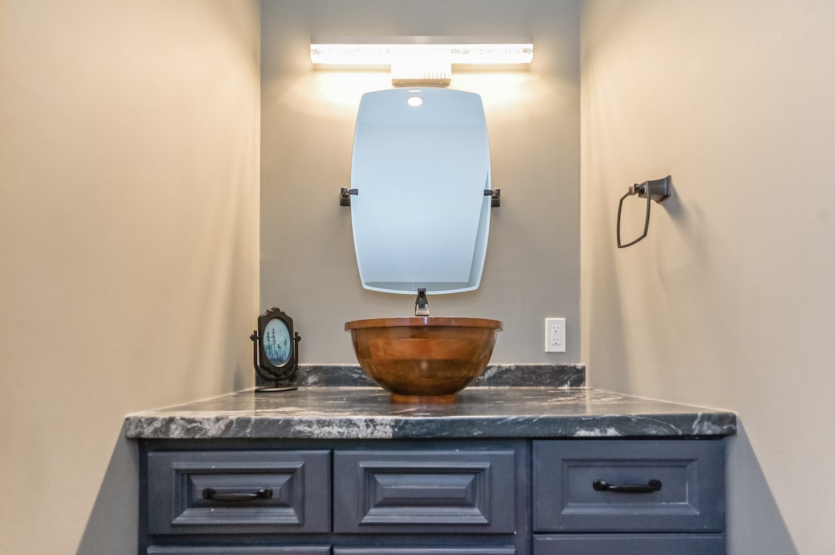 A modern bathroom vanity is showcased, featuring a sleek granite countertop and a wooden bowl sink. A round mirror is mounted above the sink, with ambient lighting highlighting the space. A wall-mounted towel holder is positioned beside the sink.