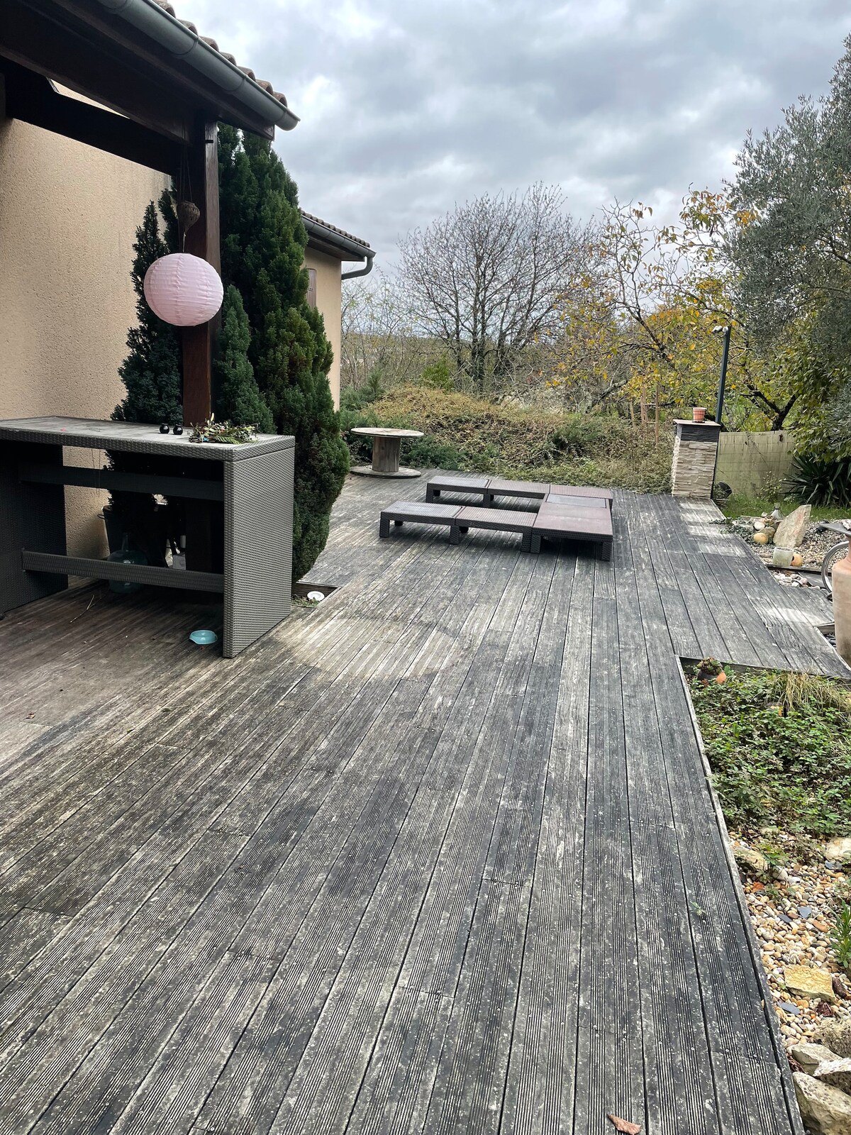An outdoor terrace features wooden decking with a simple outdoor seating arrangement. A small table and benches are visible. Lush greenery surrounds the area, creating a natural backdrop. A decorative lantern hangs nearby, adding a touch of interest to the space.