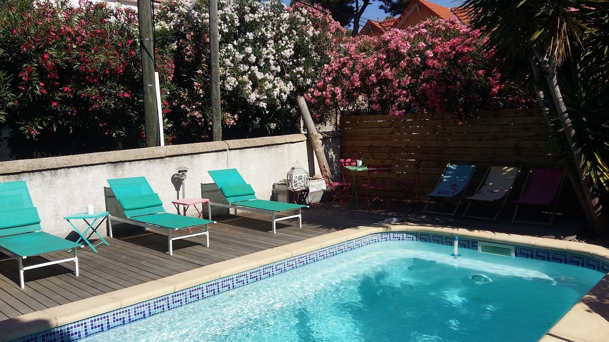 A private pool is surrounded by a wooden deck featuring lounge chairs in turquoise. Lush flowering plants create a vibrant backdrop, and a small table with chairs is positioned nearby. Sunlight reflects off the water, inviting relaxation in a tranquil outdoor setting.