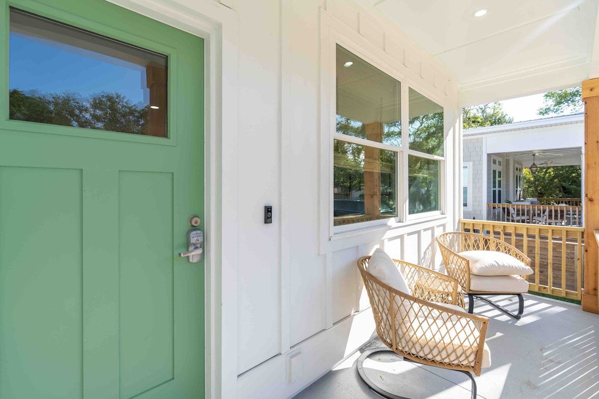 A welcoming entry features a light green front door, flanked by two stylish chairs with soft cushions. Natural light enters through large windows, illuminating the space. The porch offers a relaxing spot to enjoy the outdoor views and fresh air.