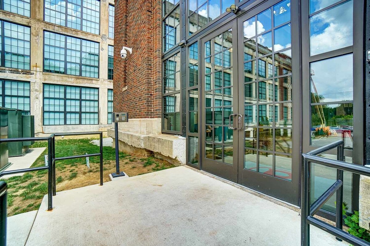 The exterior entrance is framed by large, reflective glass windows and a brick facade, showcasing a blend of historic architecture and modern design. A concrete pathway leads to the glass doors, flanked by a security camera for added safety.