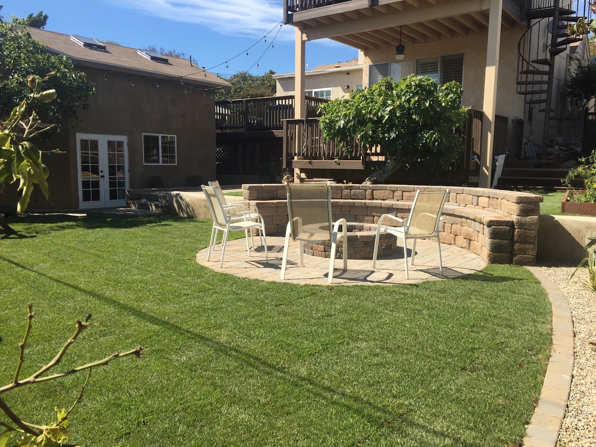 A spacious backyard features a circular stone patio with seating for six, surrounded by well-maintained grass and lush greenery. A two-story guest house is visible in the background, complete with a deck and spiral staircase. Soft overhead lights add an ambient touch to the outdoor space.