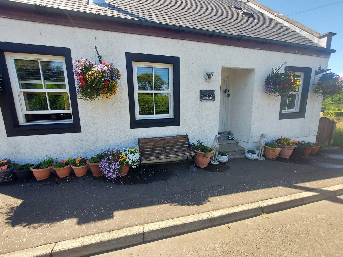 The exterior of the cottage is visible, featuring a welcoming entryway with a bench and an assortment of potted flowers in vibrant colors. Two windows adorned with floral baskets provide natural light, enhancing the charming atmosphere of the space.