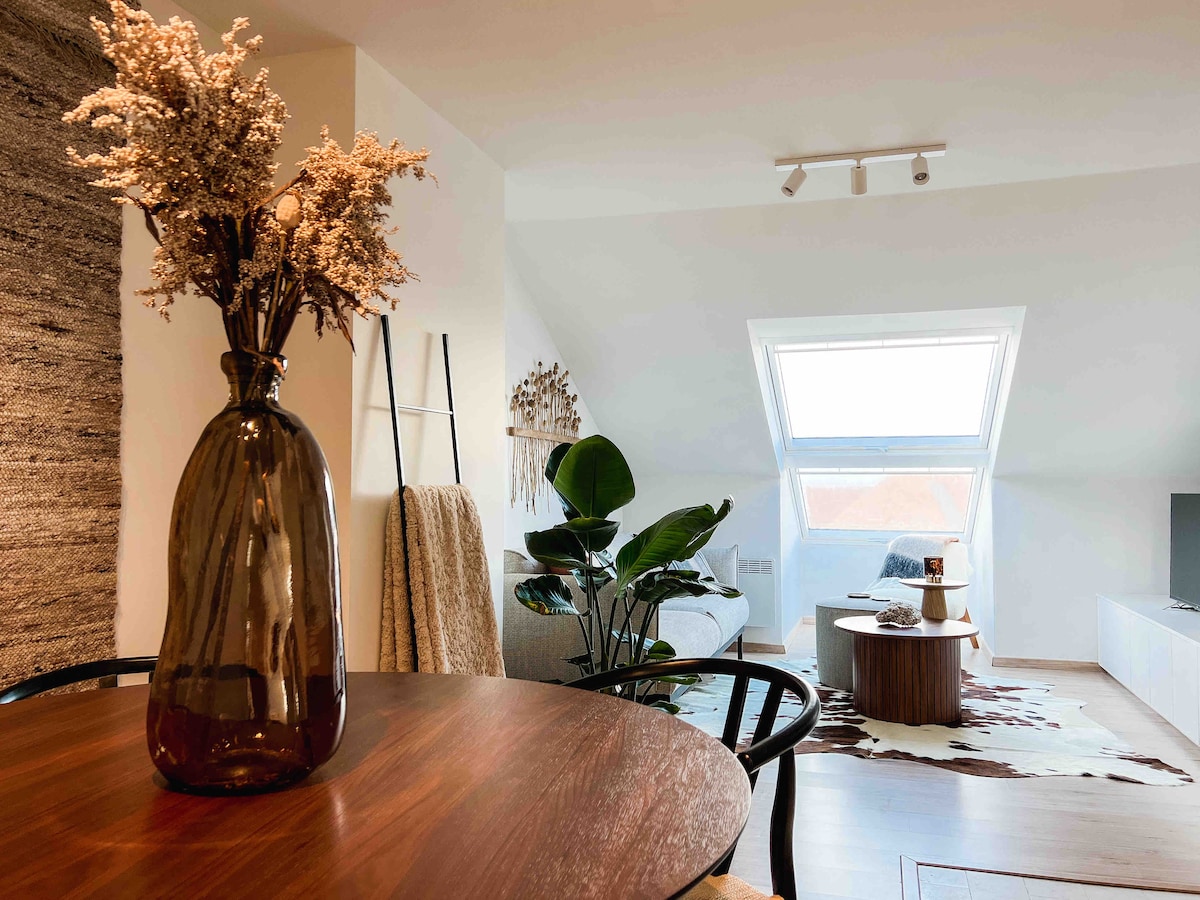 A dining area features a wooden table accompanied by a black chair. A large, brown vase holds dried flowers. In the background, a cozy living space is visible with a light-colored sofa and a large window, allowing natural light to fill the room.