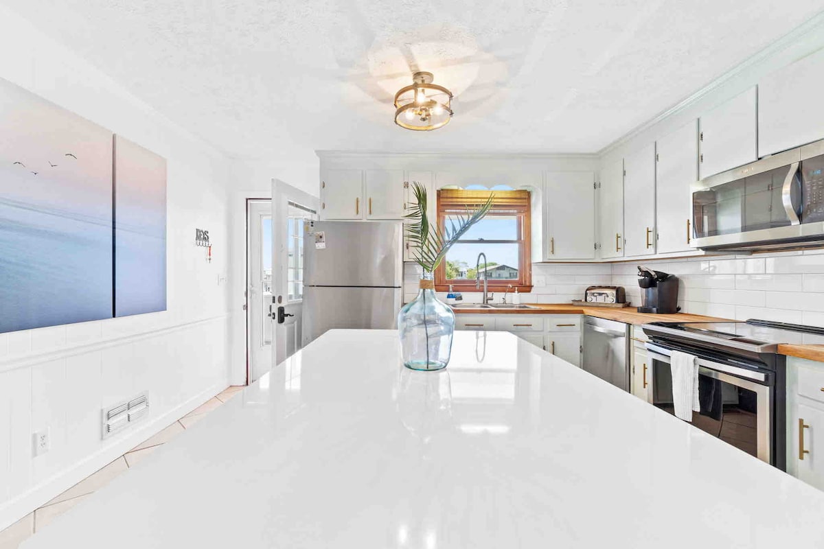 The kitchen features a spacious stone countertop island with seating for two and a modern pendant light overhead. Bright cabinetry complements stainless steel appliances, including a refrigerator and stove. Natural light fills the space through a window, offering a view of the canal.