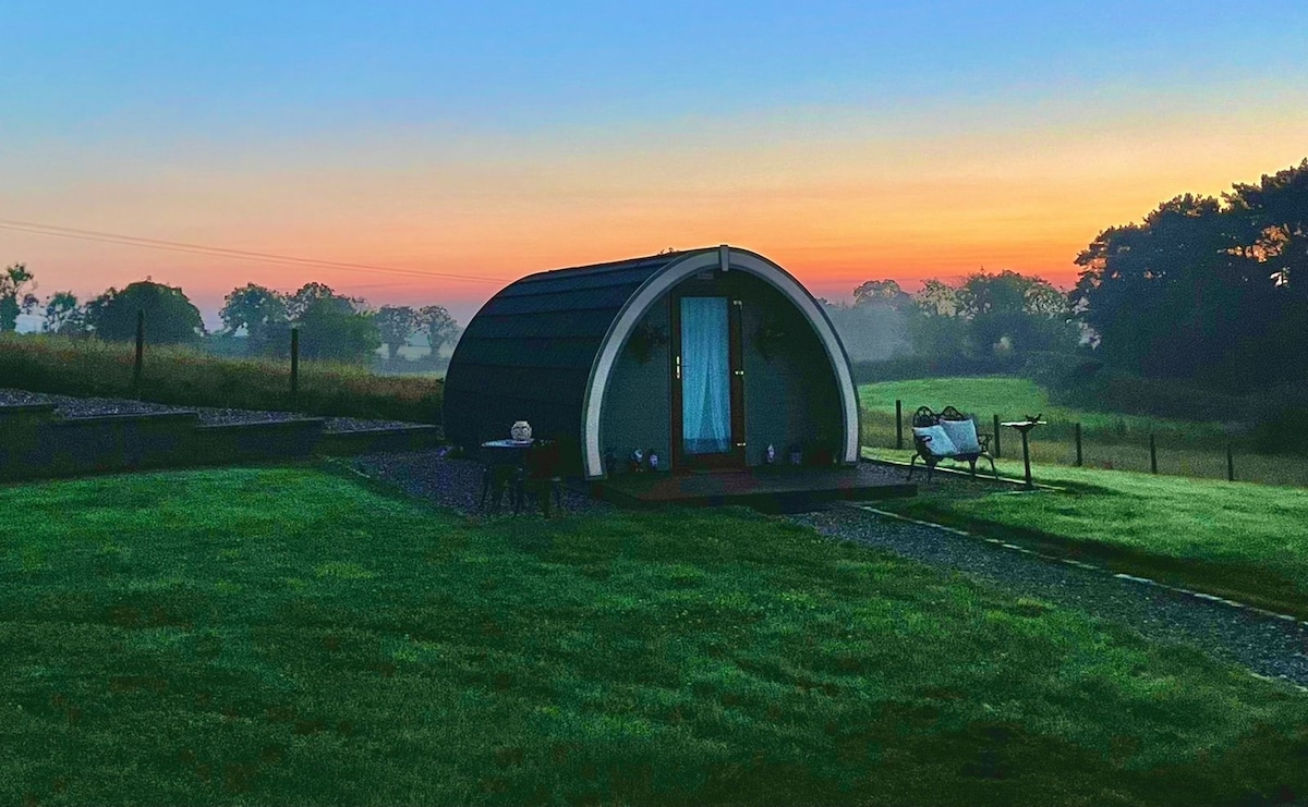 A charming glamping pod is set against a serene twilight sky, with soft hues of orange and purple blending overhead. Lush green grass surrounds the structure, while a gravel path leads to the entrance. Nearby, a seating area offers a peaceful spot to enjoy the outdoors.