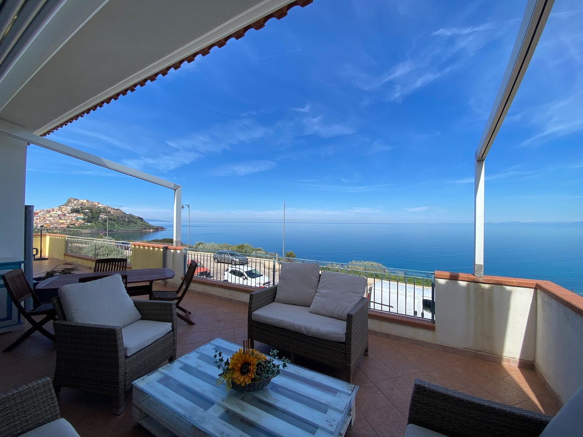 A spacious veranda offers views of the sea and distant hills, adorned with comfortable furnishings including a table and seating area. The blue sky stretches above, and sunlight illuminates the space, creating a serene outdoor setting for relaxation.