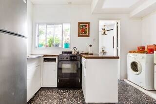 A functional kitchen features a stainless steel refrigerator and a black stove with an oven. Natural light enters through a window, illuminating the counter space which is accented by a framed artwork. A washing machine is positioned beside the kitchen area.