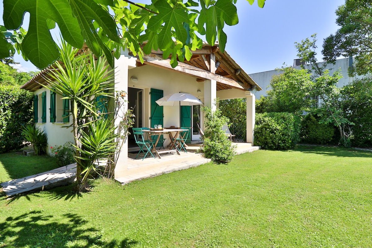 A charming cottage is surrounded by lush greenery, featuring a covered patio with a dining area. Tropical plants and manicured lawns create a serene outdoor space, while bright green shutters frame large windows that allow natural light to fill the interior.