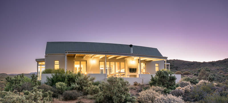 The cottage showcases a modern design with a large, wrapped veranda and panoramic views of the surrounding fynbos and mountains. Soft lighting emanates from the windows, creating a welcoming glow against the twilight sky. A tin roof complements the rustic setting.