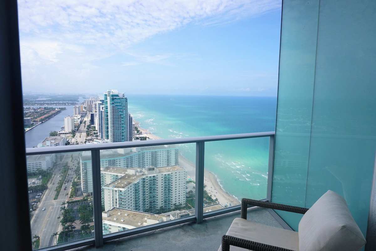 A private terrace is visible with a single armchair positioned along the glass railing. The expansive ocean view stretches out to the horizon, displaying varying shades of blue in the water. The coastline is lined with buildings, creating a striking contrast against the natural beauty.