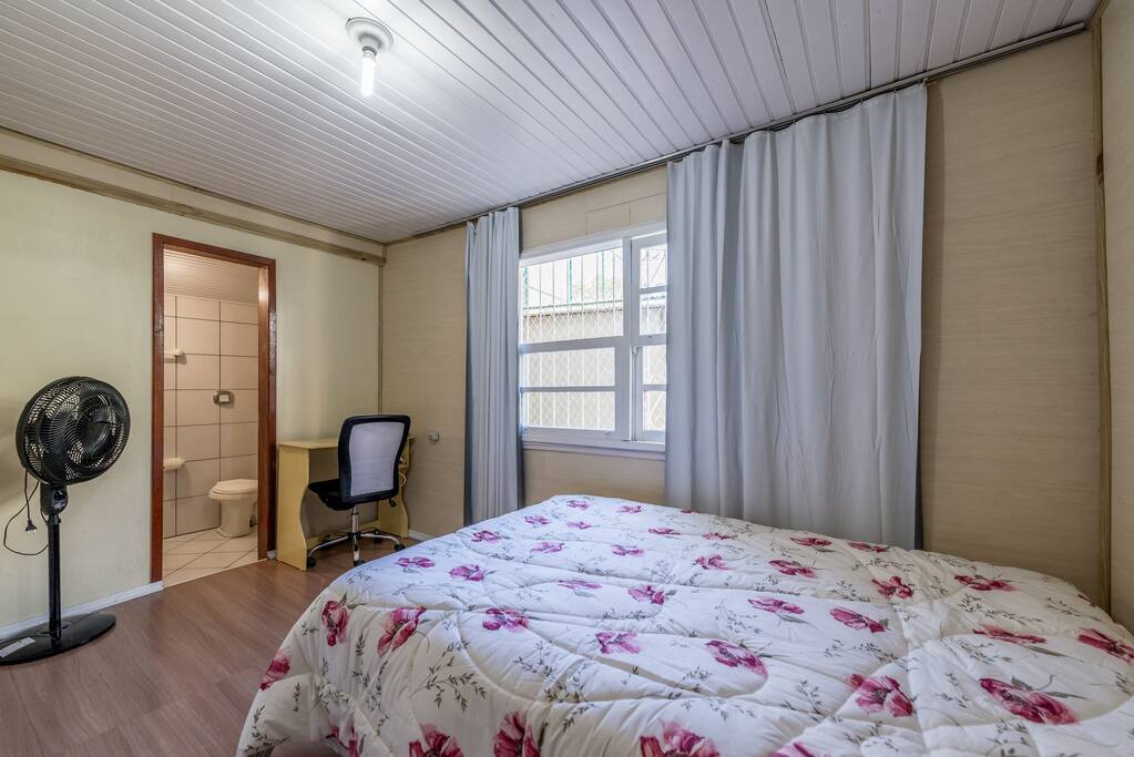 Suite ampla, c/ cama de casal, mesa e cadeira, ventilador, cortina blackout, cabideiro atrás da porta, iluminado e sol da manhã.