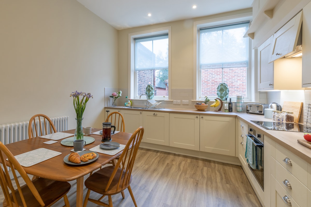 The kitchen/diner features light-colored cabinetry and large windows that allow natural light to fill the space. A wooden table with four chairs is set with plates, cutlery, and a small vase of flowers, while appliances and a variety of fruits are visible on the counters.