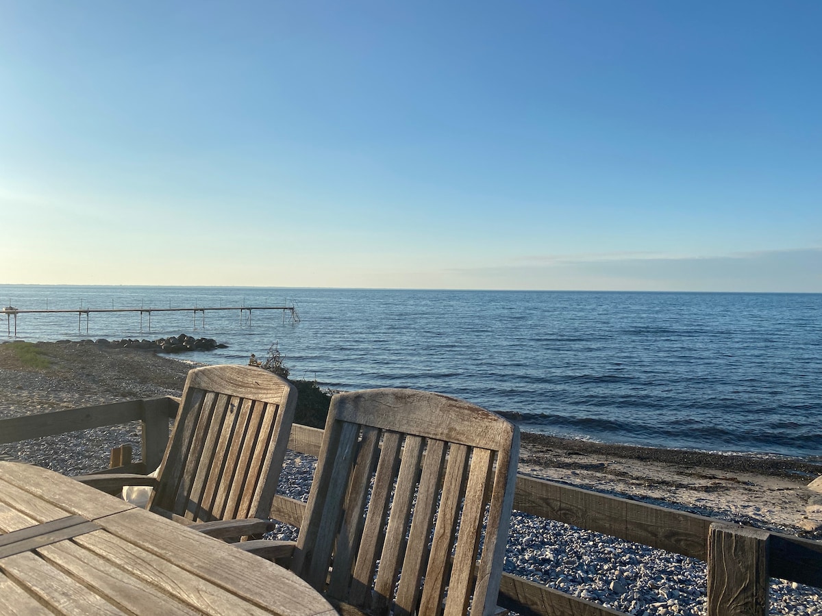 The image captures a serene coastal scene, featuring smooth calm waters that stretch towards the horizon. A wooden table with two chairs is positioned in the foreground, allowing for an unobstructed view of the ocean, where a small dock extends into the water.