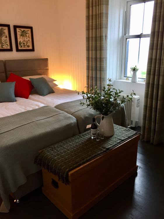 The bedroom features two single beds, configurable as a king-sized bed, adorned with decorative pillows. A wooden trunk acts as a bedside table, displaying a small vase with greenery and a bottle. Soft curtains frame the window, which offers natural light.