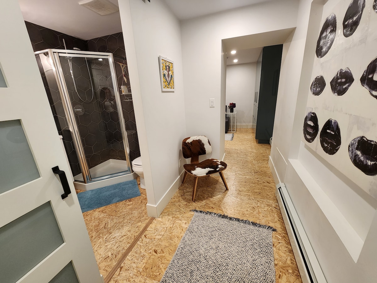 A bright hallway is visible, leading to a modern bathroom with a glass shower and light-colored walls. A stylish chair with a patterned throw sits against the wall, complemented by a textured rug. Artwork is displayed nearby, enhancing the contemporary atmosphere.