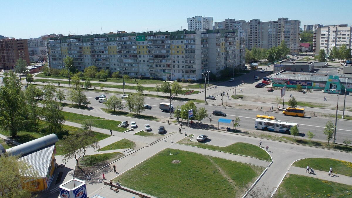 A panoramic view captures urban buildings and bustling streets. Multiple vehicles are seen traveling along the road, with pedestrians walking on the sidewalks. Green patches of grass and trees line the area, providing a contrast to the surrounding concrete structures.