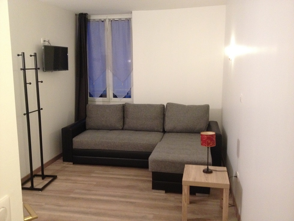 A modern living area features a gray sectional sofa positioned against a light wall. A small wooden side table with a red lamp complements the space. A coat rack stands nearby, and natural light enters through a window with sheer blue curtains.