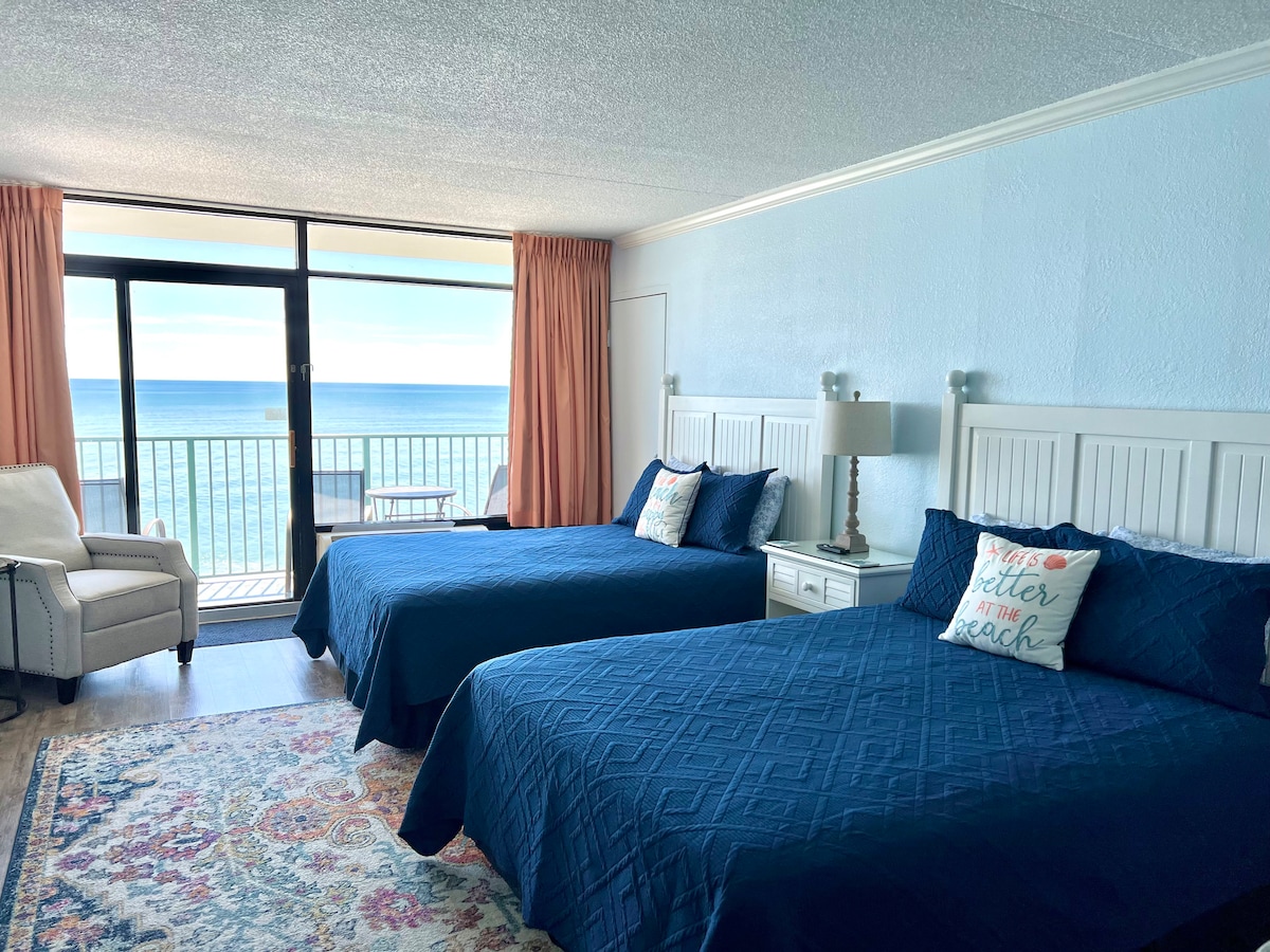 A bright and airy bedroom features two spacious beds with navy blue covers, complemented by light-colored headboards. A comfortable chair is positioned near sliding glass doors that open to a private balcony, offering views of the ocean. Soft natural light fills the room.
