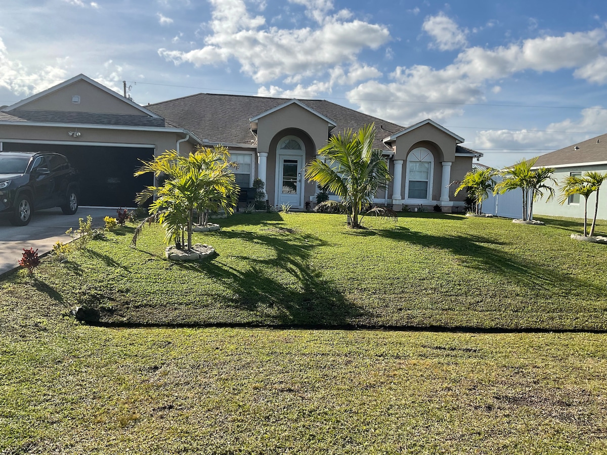 A charming house is set against a bright, partly cloudy sky. Lush palm trees and manicured grass enhance the inviting landscape. The welcoming entrance features an arched doorway, with a driveway leading to a garage on the left. Neighboring homes are visible in the background.