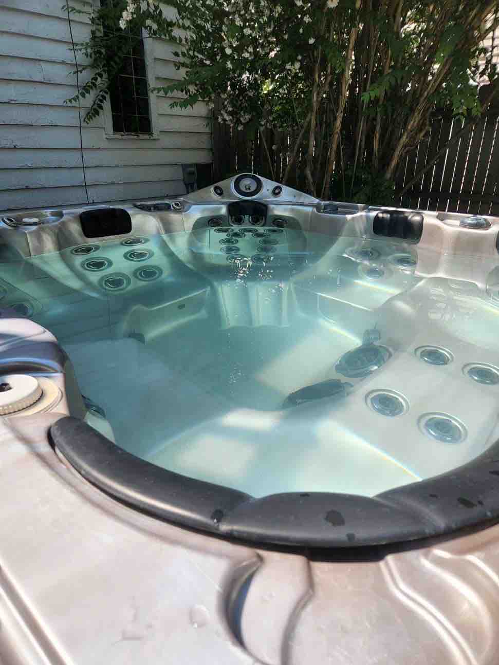 An inviting hot tub is displayed in a backyard setting, featuring numerous jets along the sides. The water appears clear and tranquil, surrounded by greenery and wooden fencing, providing a private and relaxing atmosphere.
