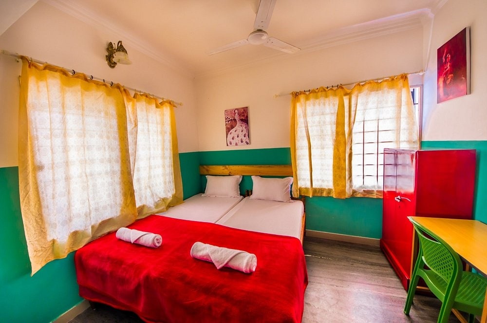 A bright room features two beds adorned with crisp linens and red throws. Soft curtains frame the windows, allowing natural light to filter in. A simple wooden desk with a green chair and a red cabinet adds a pop of color to the space.