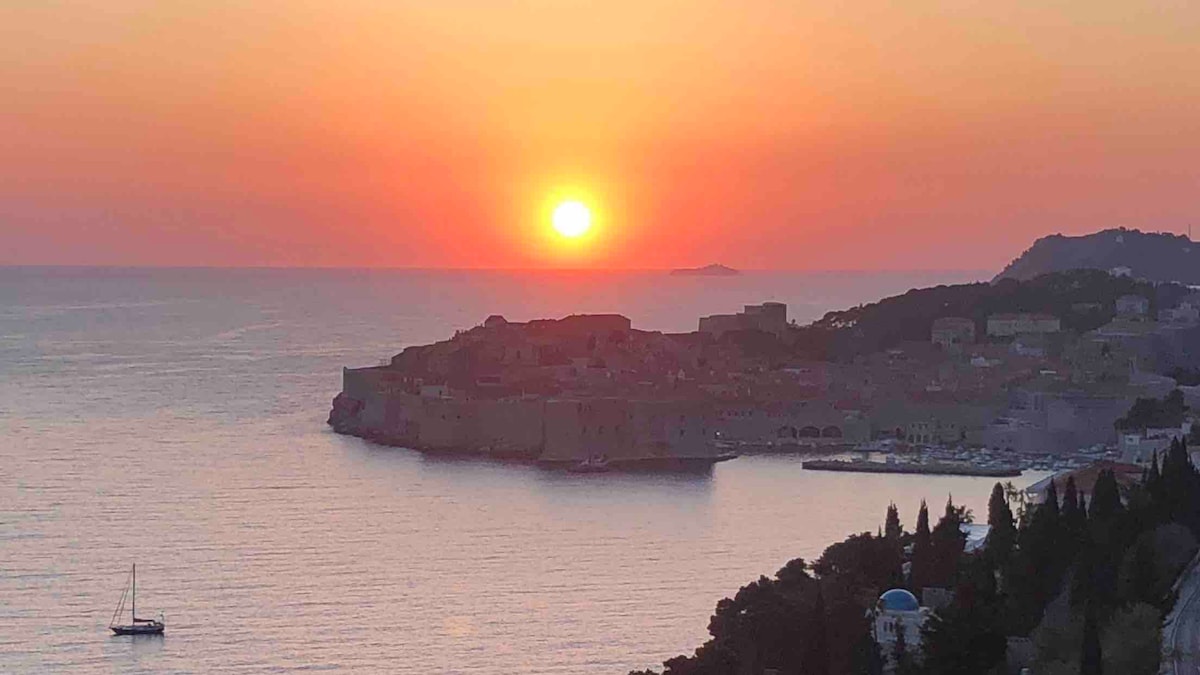 A vibrant sunset casts warm orange and pink hues over the Adriatic Sea. The silhouette of Dubrovnik’s city walls is visible against the sky, while a distant sailboat gently floats on the water. Green hills and trees frame the scene, enhancing the coastal view.