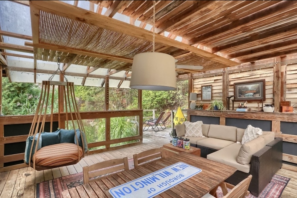 A covered outdoor space features a cozy seating area with a sectional sofa and a swing chair, surrounded by nature. Wooden panels line the walls, and a large light fixture hangs from the ceiling. A dining table sits at the center, inviting shared meals and conversation.