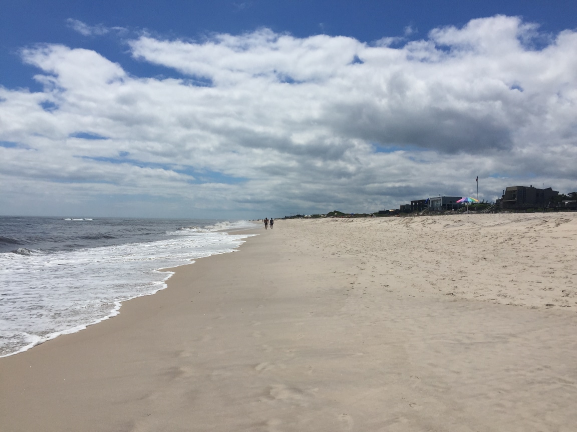 A wide sandy beach stretches under a blue sky dotted with clouds, with gentle waves lapping at the shore. In the distance, two figures walk along the water's edge, while various beachside buildings are faintly visible on the right.