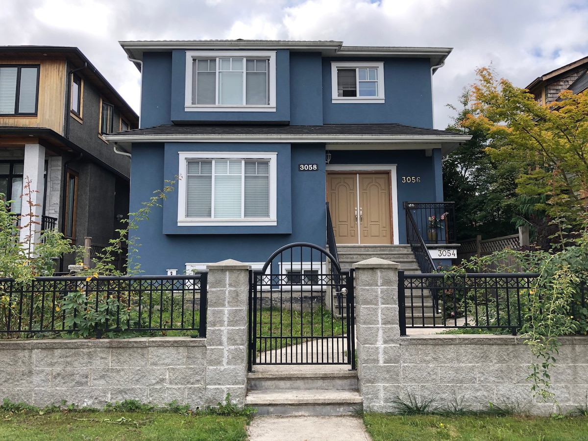 A two-story blue house is showcased, featuring a spacious front yard with green plants. A black wrought-iron gate leads to the entrance, where a set of stairs ascends to a tan door framed by light-colored window shutters. A clear sky with scattered clouds is visible overhead.